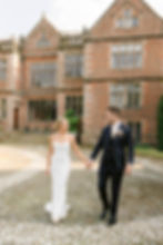 Bride in white dress and groom in dark suit holding hands, walking on cobblestone path. Background of historic brick building, both smiling.