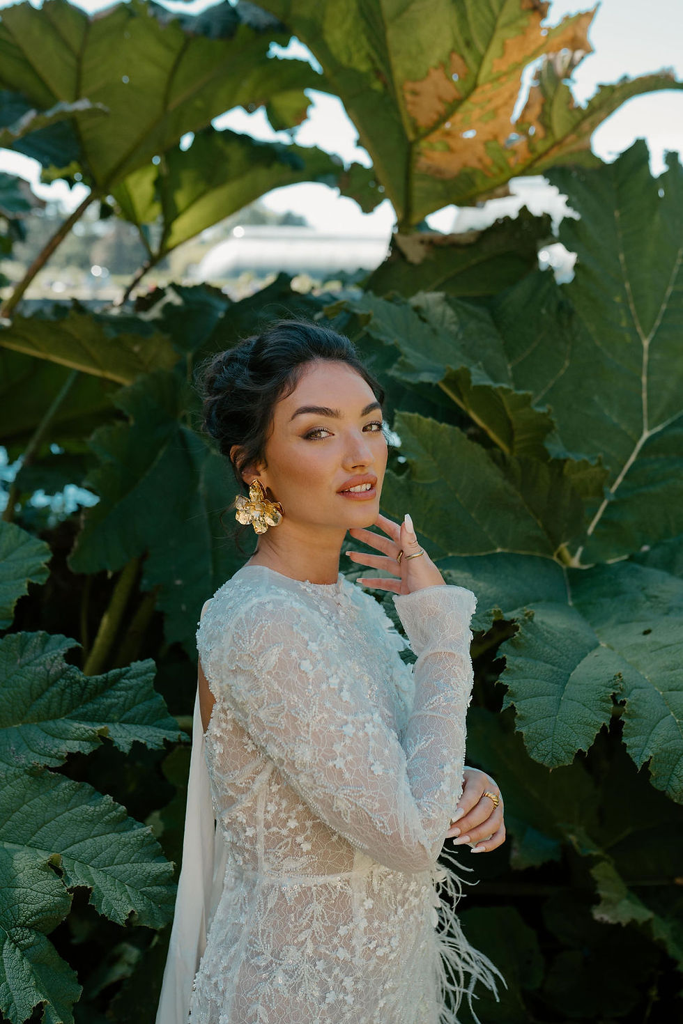 Woman in ornate white lace dress stands amid lush green foliage, wearing gold floral earrings, gazing calmly at the camera.