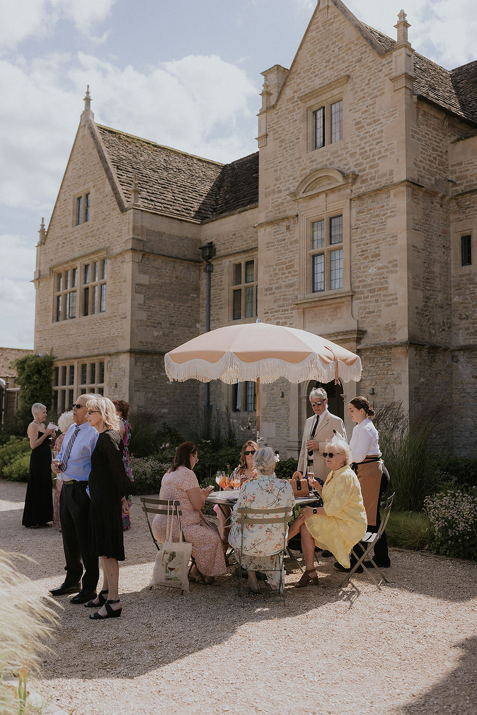 Guests mingle and sip drinks under a parasol outside a stone manor, with a warm, relaxed garden-party mood.