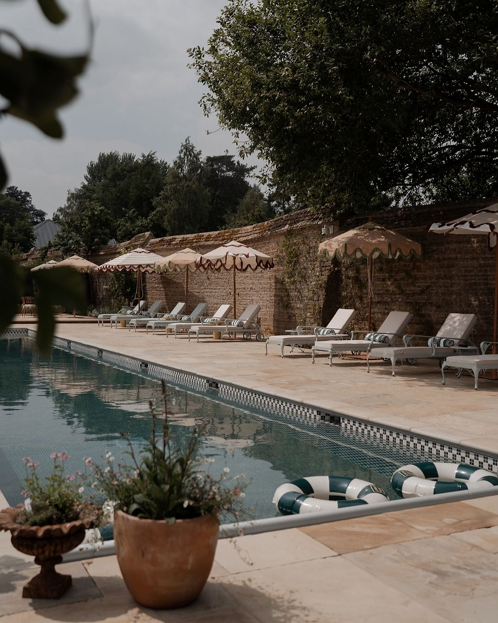Quiet outdoor pool with lounge chairs, striped umbrellas, and floating rings beside a brick wall and potted plants