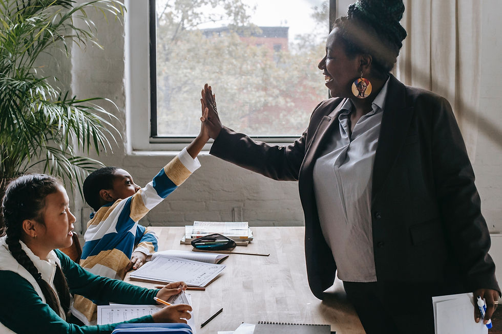 Teacher and student high-five at a table with books. Another student watches. Bright room with window and plant, fostering a positive mood.