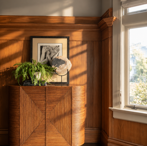 Wood-paneled corner with a fluted rattan cabinet, fern and framed art by the window