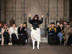 Designer in dark attire and sunglasses waves on a runway. Seated audience claps, wearing diverse fashion. Ornate iron backdrop.