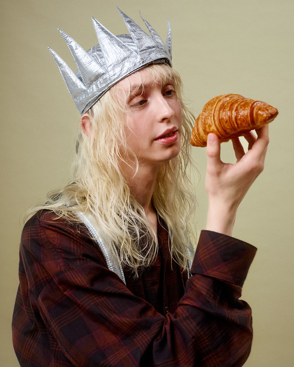 Blond person in a silver spiked crown studies a croissant against a plain beige background.