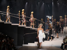 Milan Fashion Week Model in white dress leads runway, others in floral dresses behind. Dark stage with bright lights. Audience watches, some taking photos.