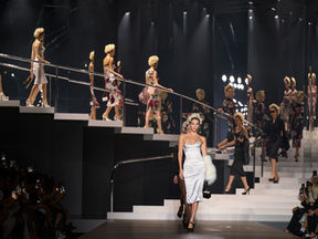 Milan Fashion Week Model in white dress leads runway, others in floral dresses behind. Dark stage with bright lights. Audience watches, some taking photos.