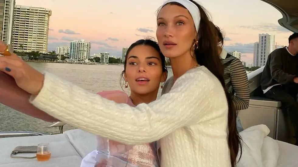 Bella Hadid and Kendall Jenner on a boat take a selfie at sunset. They're dressed in white with city buildings in the background, creating a relaxed mood.