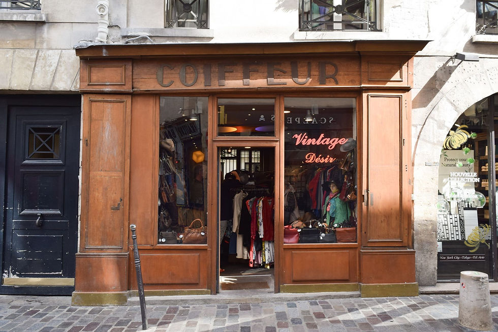 Vintage storefront with wooden façade. Neon sign reads "Vintage Désir." Clothing and accessories visible inside. Cobblestone street in Paris.