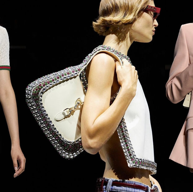 Models in Gucci outfits; one wears white knit with red-green accents, another holds a jeweled bag, and a third wears a pink suit. Stylish vibe.