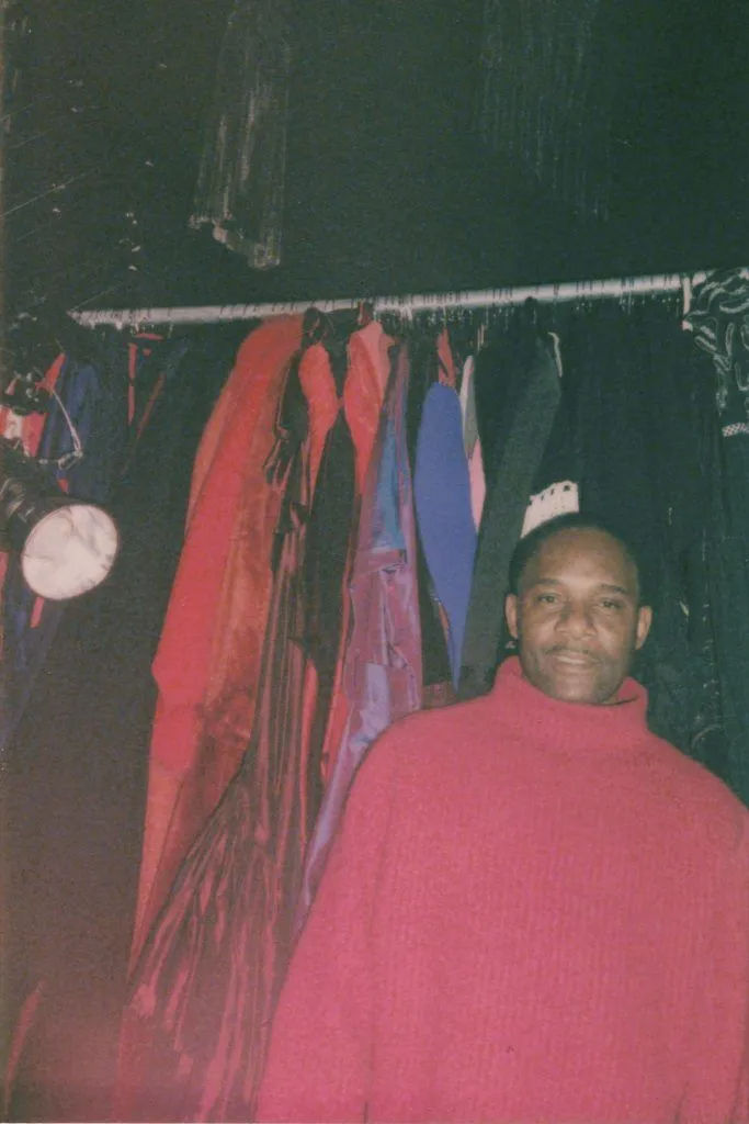 A person in a red sweater stands beside a colorful rack of clothes in a dimly lit room, creating a cozy, casual atmosphere.