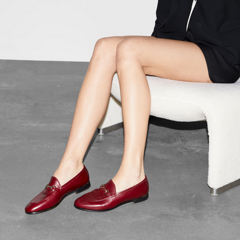 Person in a black outfit sits on a white chair, wearing red Gucci loafers on a gray floor. Focus is on the shoes. Minimalistic setting.