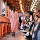 Model in pink plaid coat walks runway on a boat. Audience seated on right, taking photos. Orange metal railings and lights above.