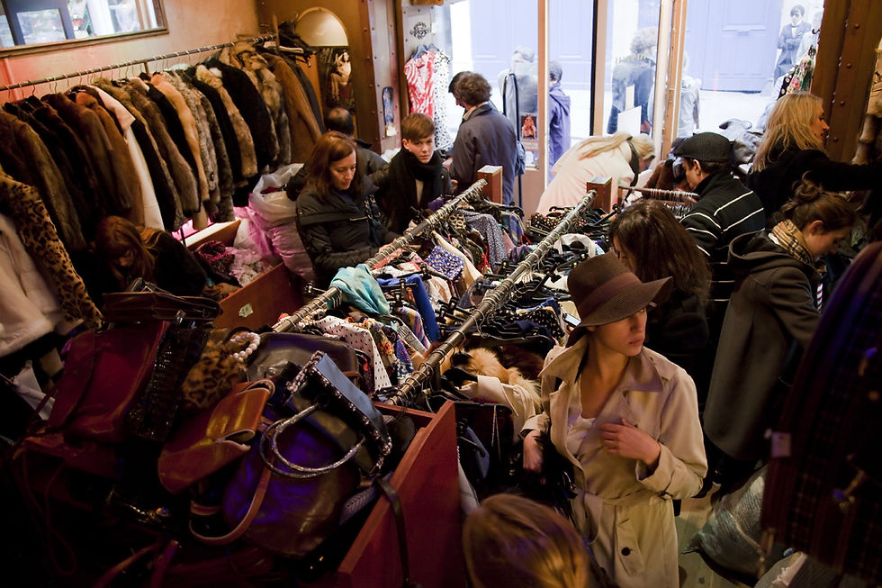 People browse racks of colorful vintage clothes in a crowded shop with fur coats, hats, and bags. The mood is busy and lively.