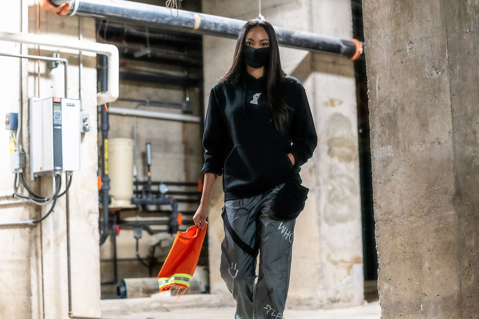 Woman in black hoodie and mask walks in industrial setting, holding an orange cloth. Grey pants with text. Dull, concrete background.