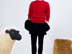 Young man in a red sweater and patterned cone hat stands in a white studio beside a sheep cutout and straw bale, looking pensive.