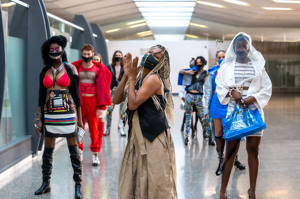 Fashion models in vibrant outfits and masks walk confidently in a modern, well-lit hallway. One claps enthusiastically in the foreground.