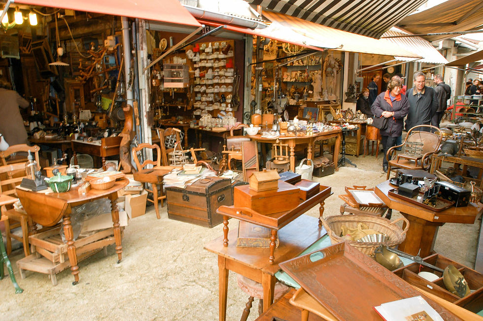 Antique market in Paris with wooden furniture, vintage items, and decor. People browsing under a striped awning. Warm tones and a busy atmosphere.