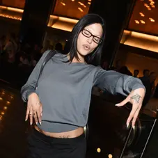 Woman in a gray sweater strikes a pose with arms outstretched, eyes closed. Dark interior with glowing lights and people in background.