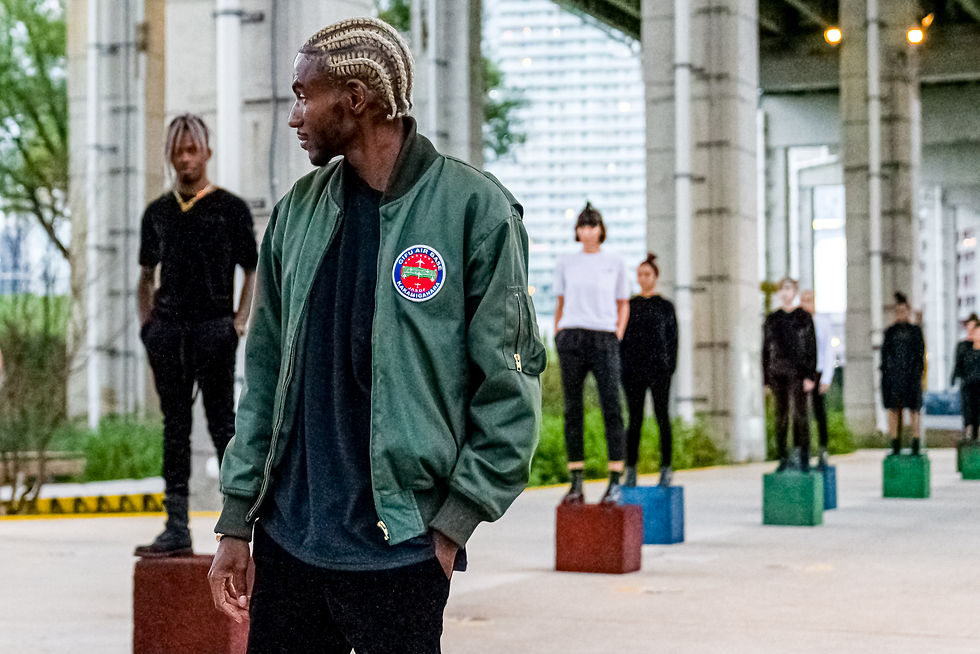 Person with braided hair in green jacket stands turned away; others in black stand on colored blocks under a bridge. Urban vibe.