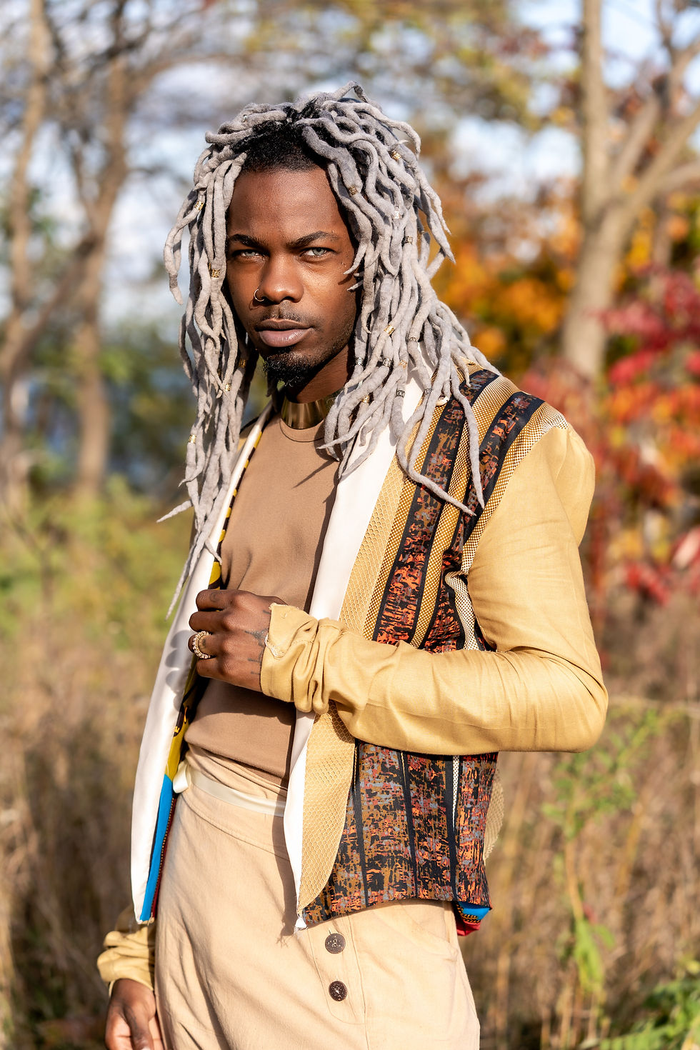 Person with gray dreadlocks in a colorful jacket and beige pants stands outdoors, surrounded by trees with autumn leaves, looking confident.