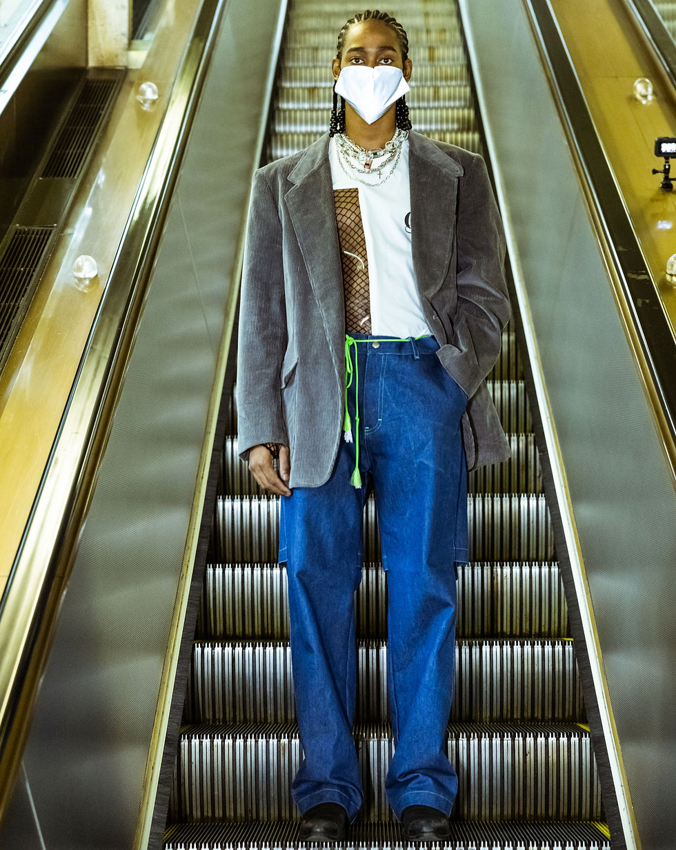 Person in a mask and gray blazer stands on an escalator in a well-lit setting, wearing a white shirt, blue jeans, and layered necklaces.