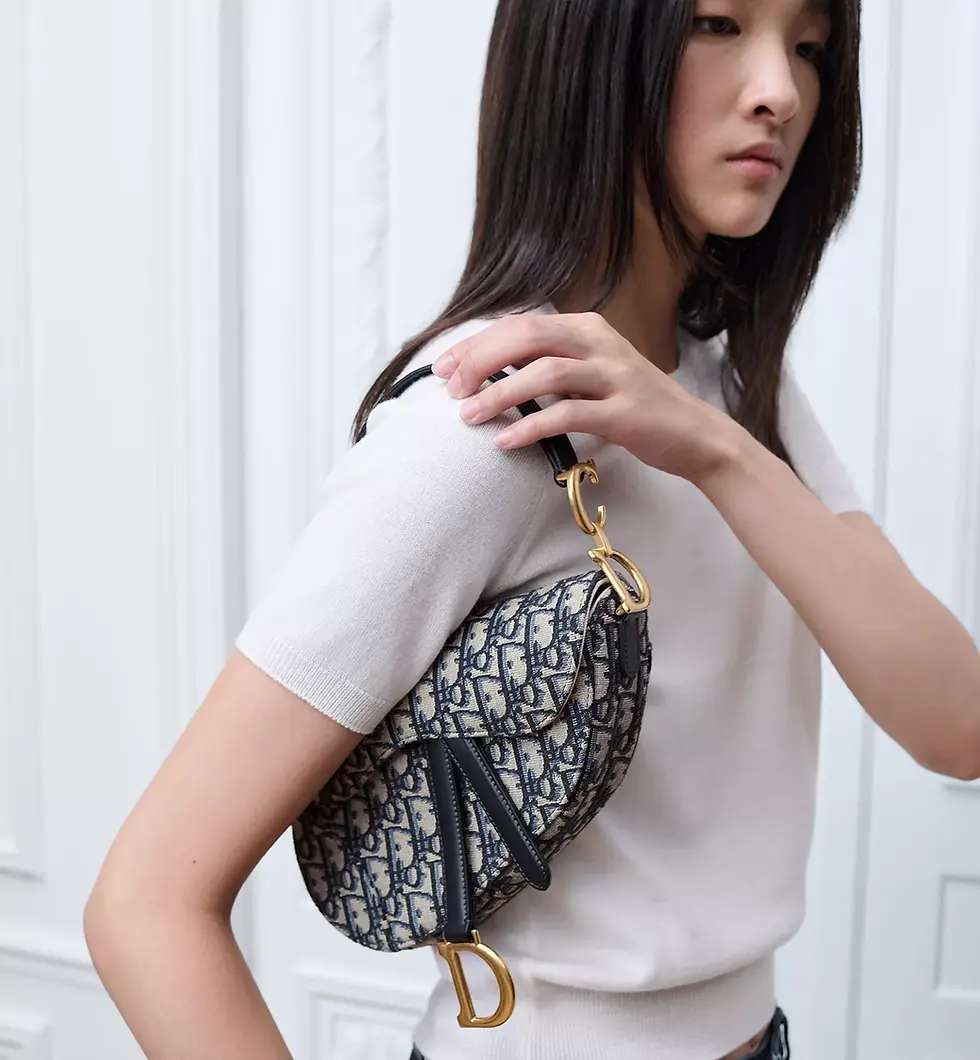 Woman in a black outfit holds three Dior Saddle bags: blue patterned with text, black leather, and white leather, each with gold detailing.