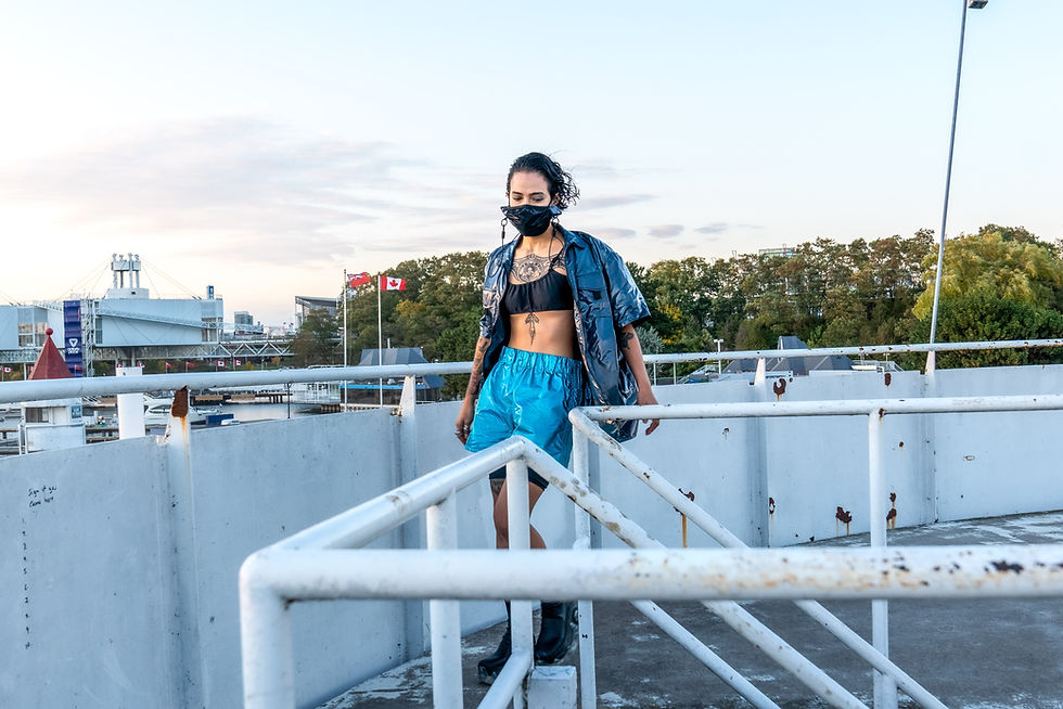 Woman in blue outfit and mask walks on a rooftop with rusty rails. Canadian flag waves in the distance against a cloudy sky.