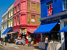 Colorful London street scene with vibrant red and blue buildings featuring shops. People browse displays under blue awnings. Union Jack flag visible.