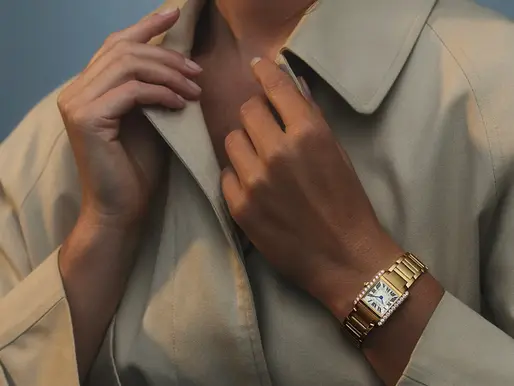 Close-up of a person adjusting a beige coat collar, wearing a gold Cartier Tank watch with a white face. Soft lighting creates a calm mood.