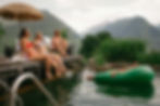3 Frauen sitzen lässig am Alpine Beach Club beim BergBaur, lassen die Füße in den Naturbadeteich baumeln und geniessen die Aussicht und den Sommertag. Ein Mann liegt entspannt auf einer aufblasbaren Avocado und lässt sich treiben,
