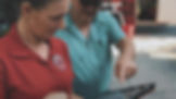 Two people examine a tablet outdoors. The woman in a red shirt with a logo looks focused, while the man in sunglasses points at the screen.