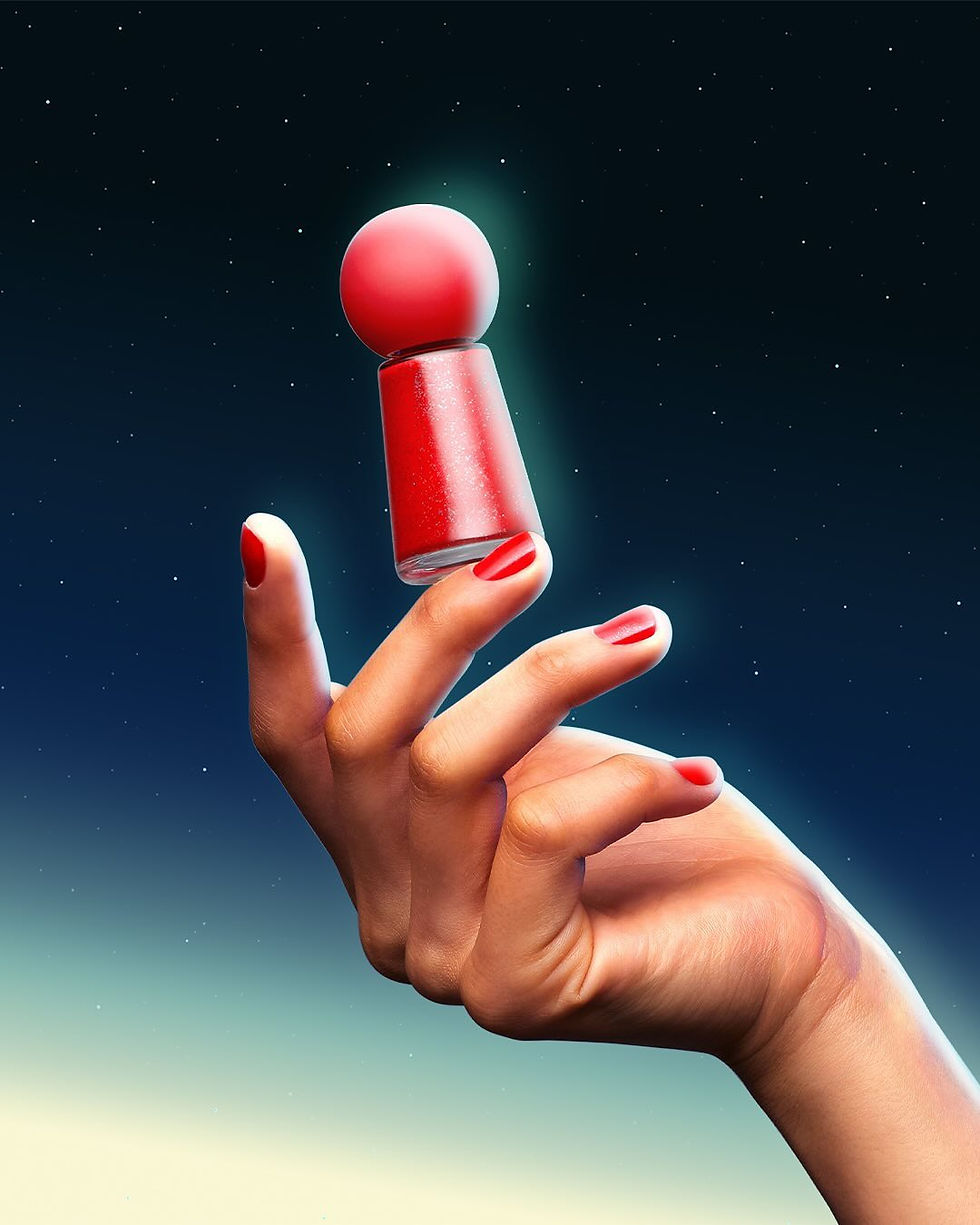 Uma mão com unhas compridas e perfeitamente esmaltadas em vermelho vibrante segura, entre os dedos indicador e médio, um frasco de esmalte vermelho cintilante. O frasco tem uma distinta tampa esférica vermelha e está posicionado contra um fundo que se assemelha a um céu noturno estrelado, com um gradiente de azul escuro para um azul-claro no horizonte.