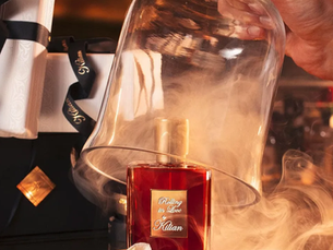 A imagem mostra um frasco do perfume Rolling in Love, da marca de luxo Kilian Paris. A fragrância, de cor vermelha, está sobre uma bandeja dourada com uma flor branca ao lado, enquanto uma cúpula de vidro é levantada, liberando uma fumaça que envolve a cena. O fundo desfocado sugere um ambiente de luxo, com caixas de presente da marca.