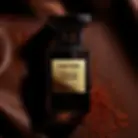 Fotografia de estúdio do perfume Tuscan Leather da marca Tom Ford. A imagem mostra o frasco de perfume preto fosco e pesado, com uma placa dourada no centro que exibe o nome da marca e da fragrância. O frasco está posicionado sobre uma superfície de couro marrom-escuro, cercado por fios de açafrão, que são uma nota chave na fragrância. A iluminação é dramática, destacando a textura do couro e do frasco.
