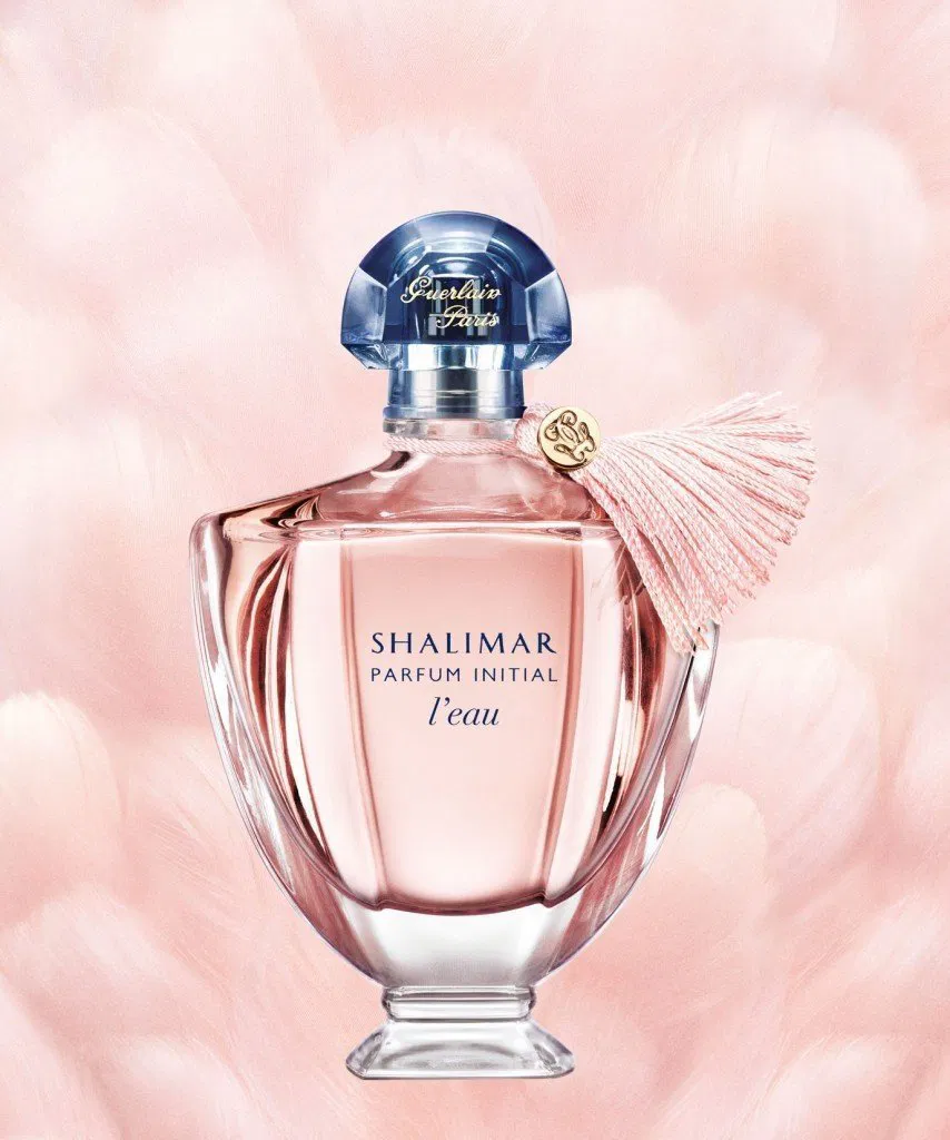 Frasco de Shalimar Parfum Initial L'eau com líquido em tom rosa pálido e um pompom (tassel) de seda rosa preso ao gargalo. O fundo é suave e rosado com texturas que lembram penas ou pétalas.