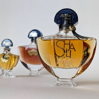 Três frascos de perfume Shalimar de diferentes tamanhos e épocas dispostos em profundidade. O frasco frontal é grande, com o nome 'SHALIMAR' em destaque no vidro e tampa azul com laço de couro preto.