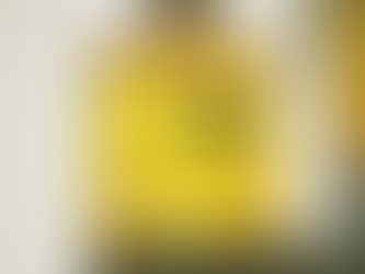 Close-up de um frasco cilíndrico de vidro transparente sendo enchido por uma máquina industrial. O frasco contém um líquido amarelo vibrante com algumas bolhas visíveis. No vidro, está estampado em preto o texto: 'ONITSUKA TIGER ONE - EAU DE PARFUM - MADE IN FRANCE'. O fundo é desfocado, sugerindo um ambiente de fábrica ou laboratório.