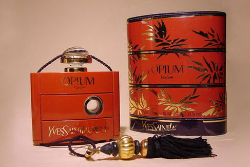 Frasco vintage do perfume Opium de Yves Saint Laurent, lançado em 1977. O frasco vermelho com detalhes dourados e formato retangular traz uma alça com cordão preto e dourado, acompanhado da embalagem cilíndrica decorada com folhas douradas sobre fundo vermelho. Ícone da perfumaria, o design evoca luxo e exotismo.
