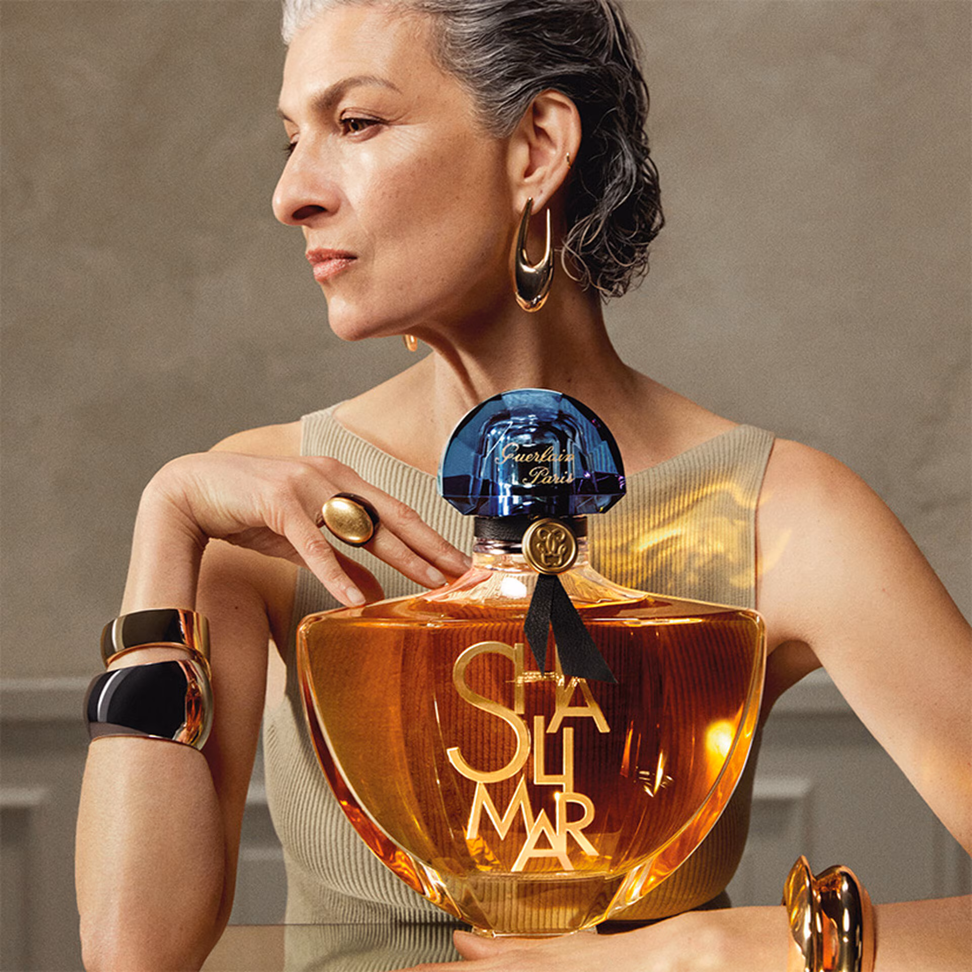 Mulher madura de cabelos grisalhos presos e joias douradas imponentes, segurando um frasco grande de perfume Shalimar contra o peito. O frasco exibe o líquido âmbar e a tampa azul clássica.