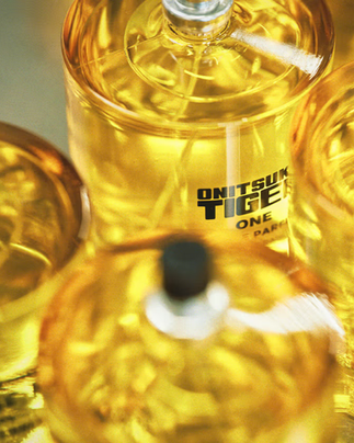 Visão superior artística e com foco suave de vários frascos de perfume agrupados. A luz reflete no vidro e no líquido amarelo-ouro, criando uma estética brilhante e etérea. O logotipo preto 'ONITSUKA TIGER ONE' é parcialmente visível no frasco central