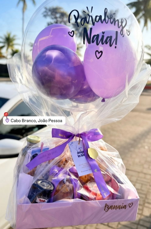 Presente em caixa rosa e roxa com balão transparente dizendo Parabéns, Naia!, em rua ensolarada com palmeiras.
