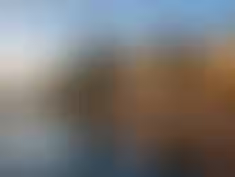 Two-story large home with floor-to-ceiling windows on both levels. Home sits on the waterfront with a ramp leading to a dock with a sailboat. It's sunset in Washington. Four large trees to the left of the house.