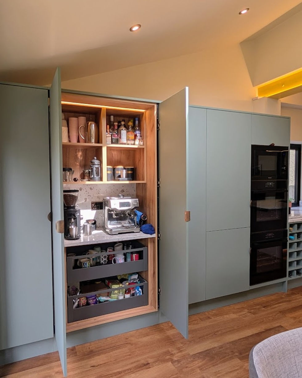 Open kitchen cabinet with coffee station, espresso machine, mugs, bottles and drawers; warm wood, sage doors, modern tidy interior