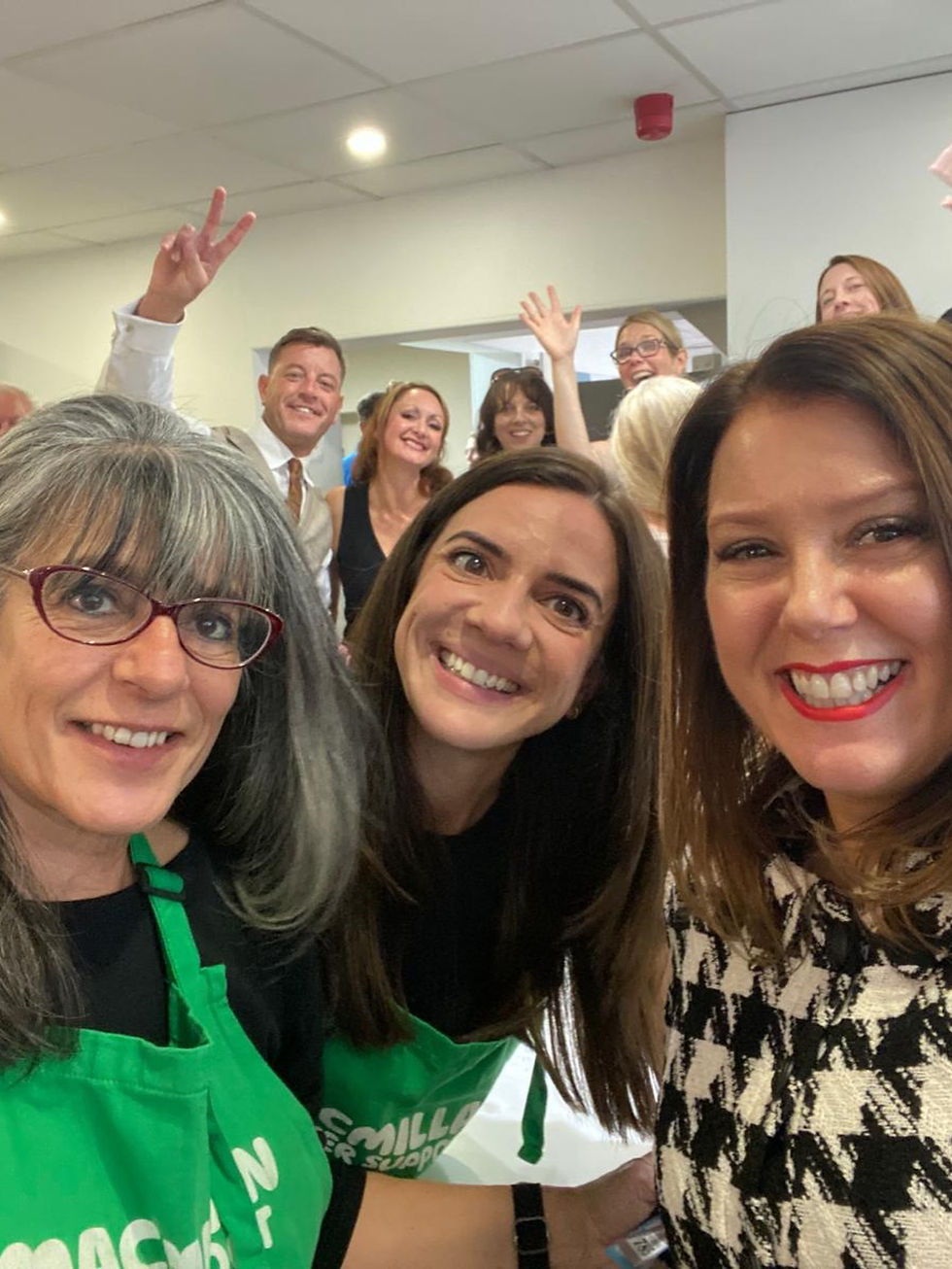 Smiling coworkers pose for a selfie indoors; one man flashes a peace sign, and two women in green aprons fill the foreground.