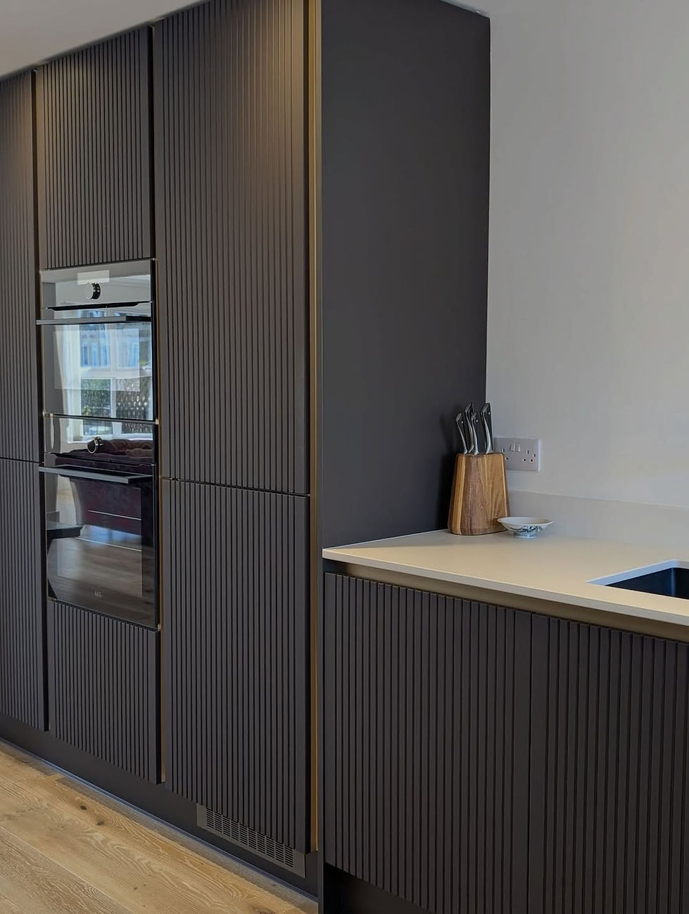 Modern kitchen with dark ribbed cabinets, built-in oven, white countertop, and knife block beside a sink.