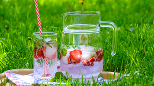 Strawberry mint iced drinks in a glass pitcher and tall glass with striped straw on a tray in bright green grass.