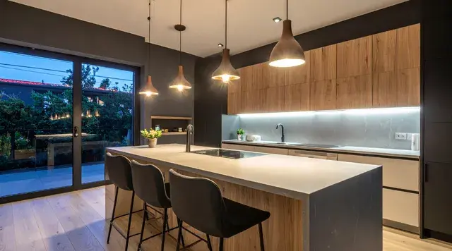 Modern kitchen with wooden cabinets, pendant lights over an island, bar stools, and a large window. The mood is warm and inviting.