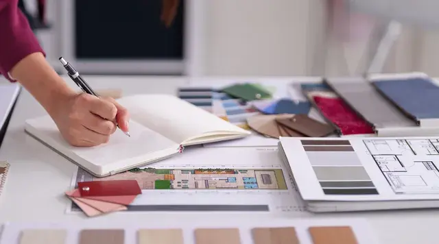A person writes in a notebook on a desk with architectural plans, color swatches, and fabric samples. The setting is bright and organized.