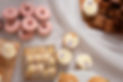 A photograph of a selection of baked goods created by Bake My Cake based in the Scottish Borders. The photograph shows iced cupcakes, macrons, baked doughnuts and brownies, all artistically laid out on a table covered in white tulle.