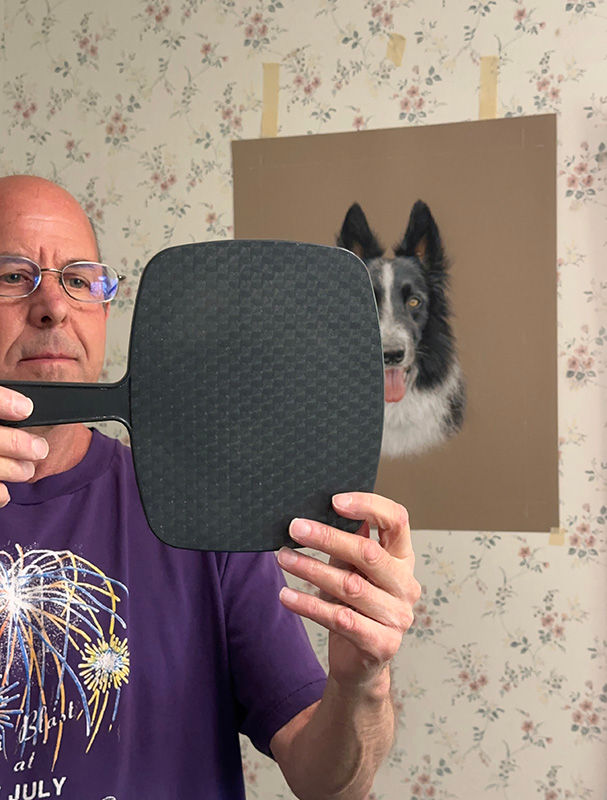 Kevin looking at his portrait of a Border Collie in a hand-mirror in his studio.